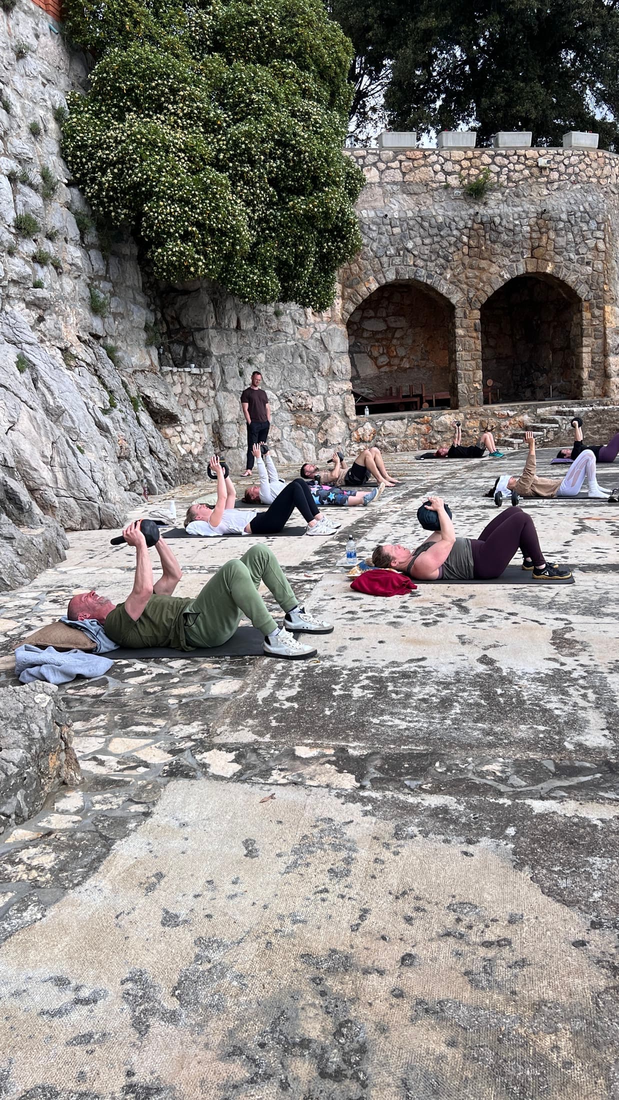 A small group on mats in a stone courtyard, working with kettlebells while a facilitator observes