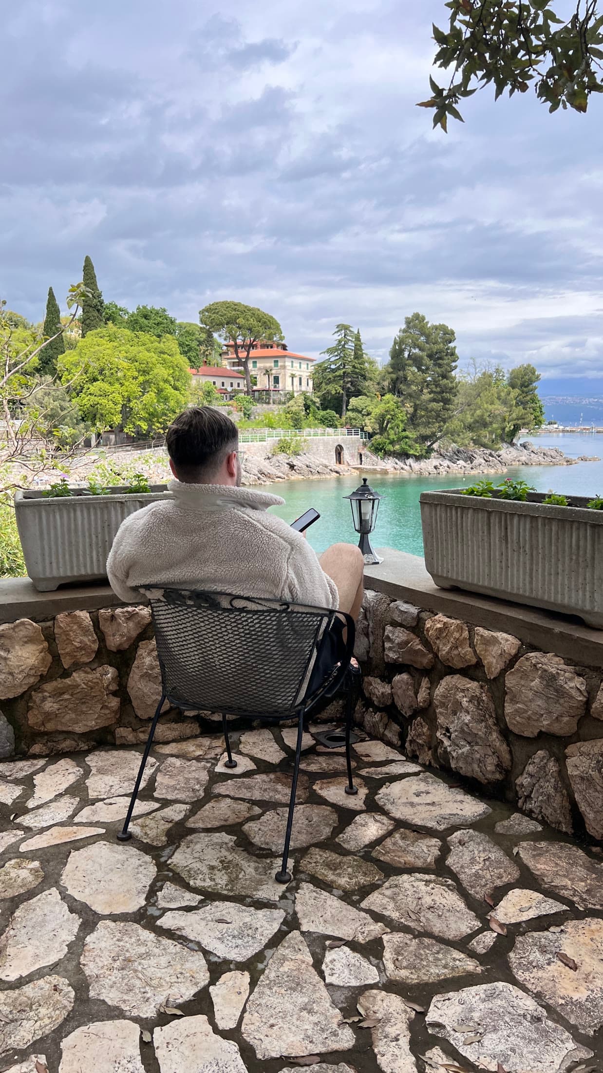 A person seated on a stone terrace, looking out over water toward a wooded shoreline