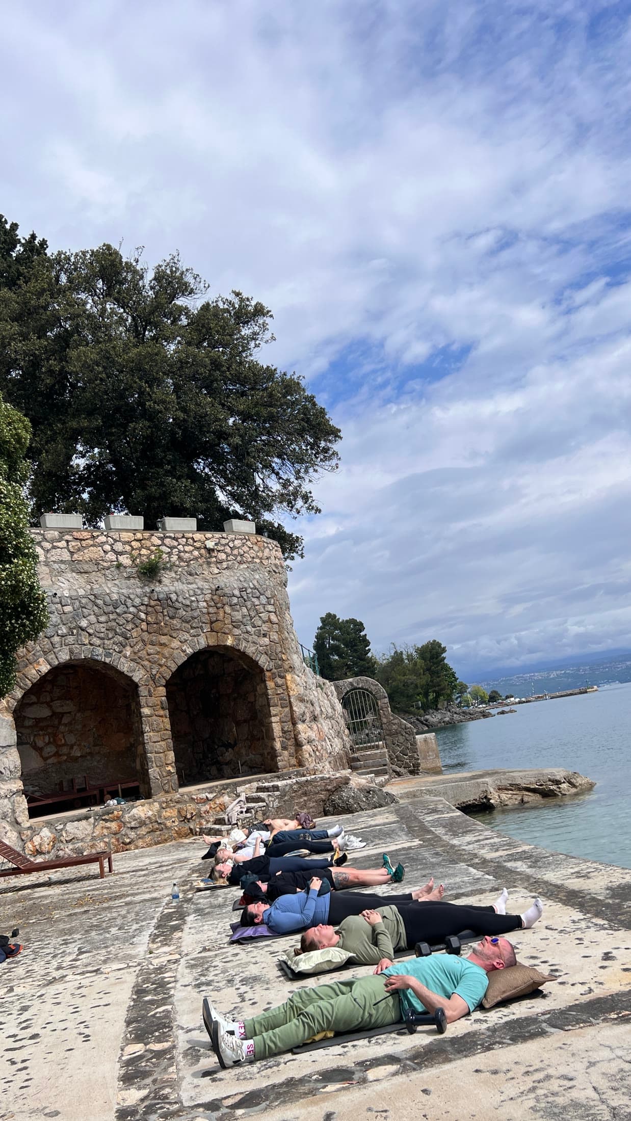 Participants lying in a line on a stone terrace beside the water, a historic arched wall to one side