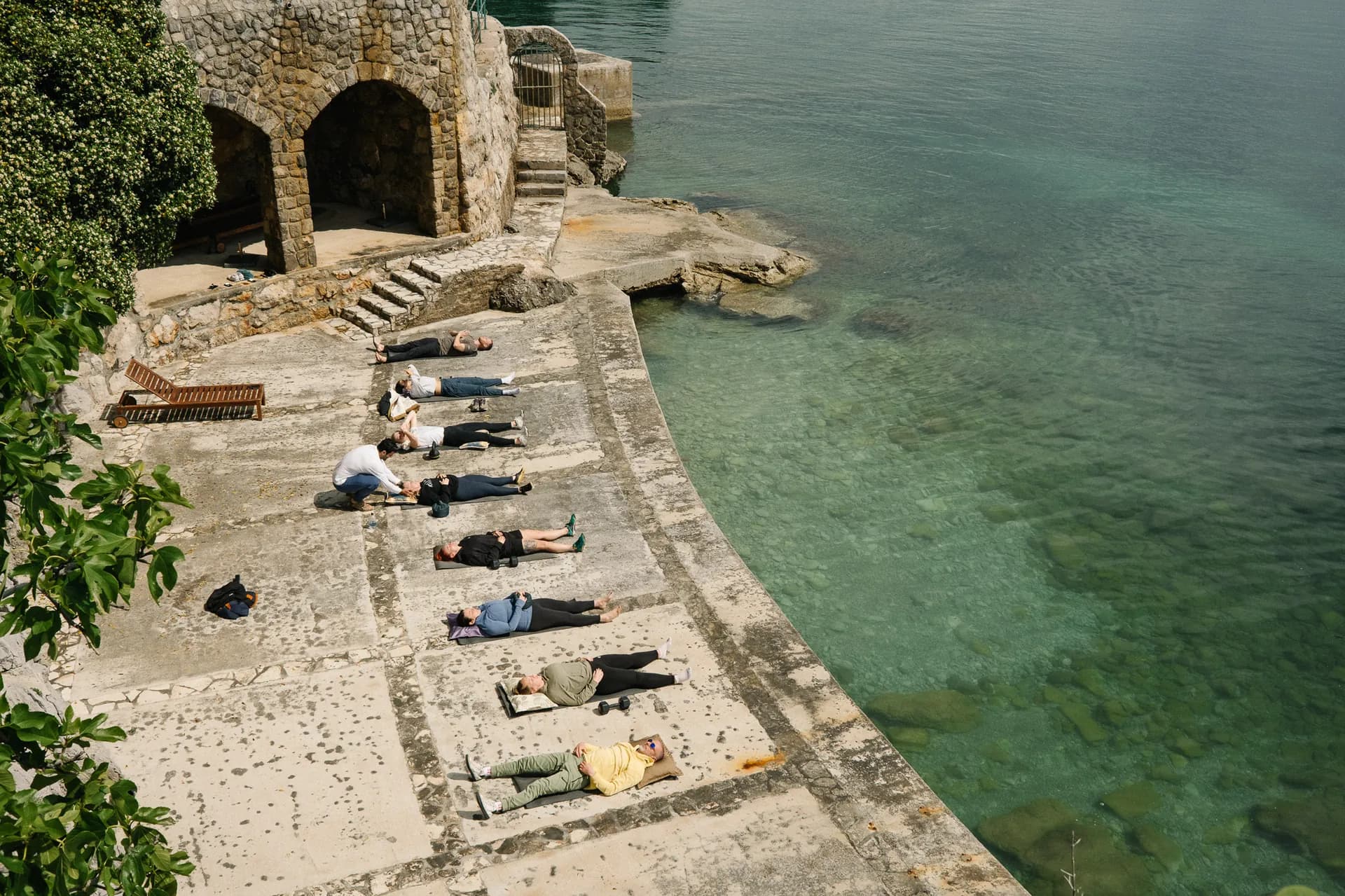 Retreat participants lying on a stone pier beside clear water during a guided session